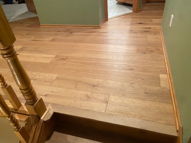 Light tan hardwood plank flooring installed in a hallway with a wooden stair railing and doorways to adjacent rooms.