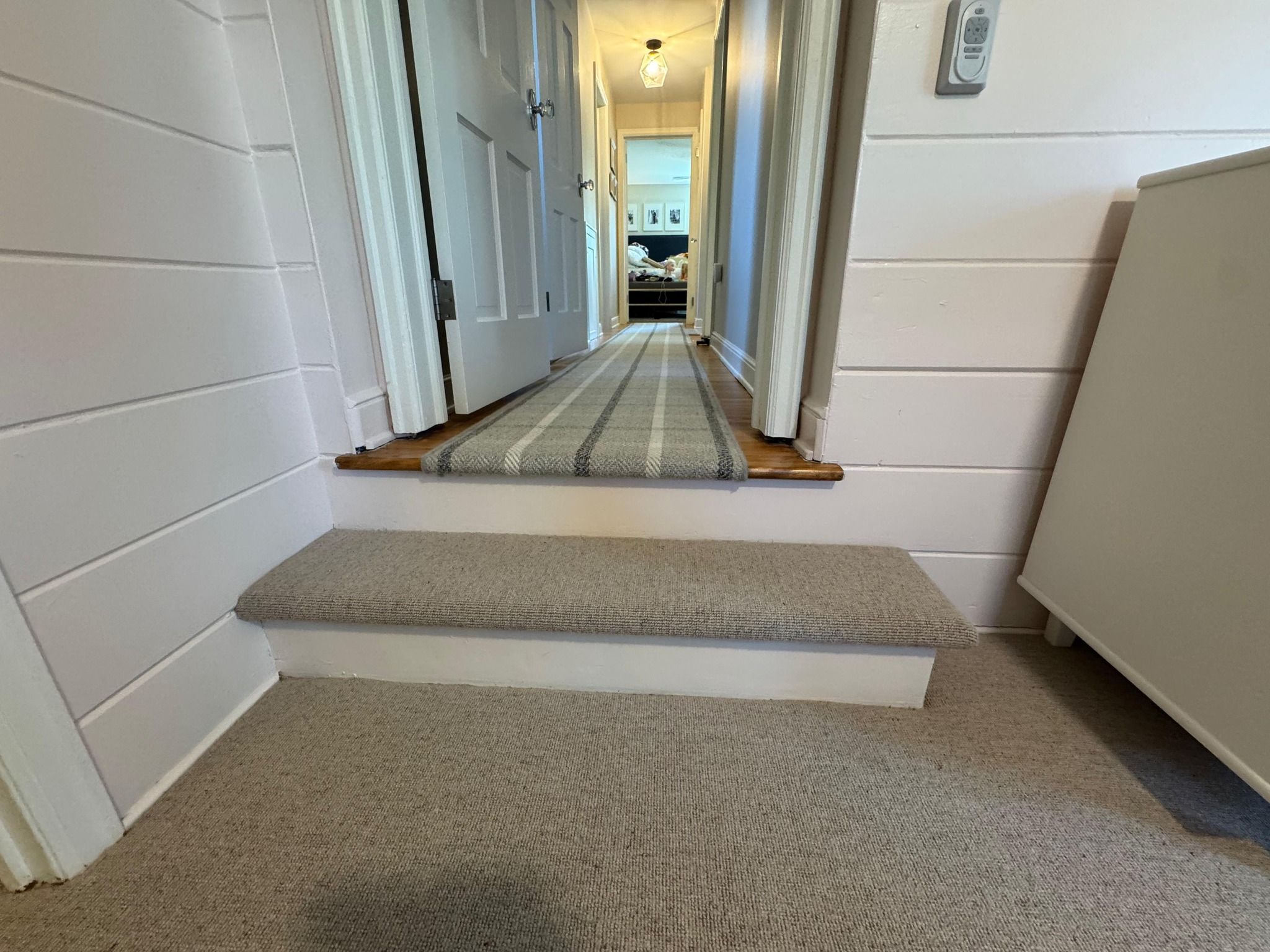 Hallway and stairs with tan wool carpet, stair runner with striped pattern, and white wall paneling.