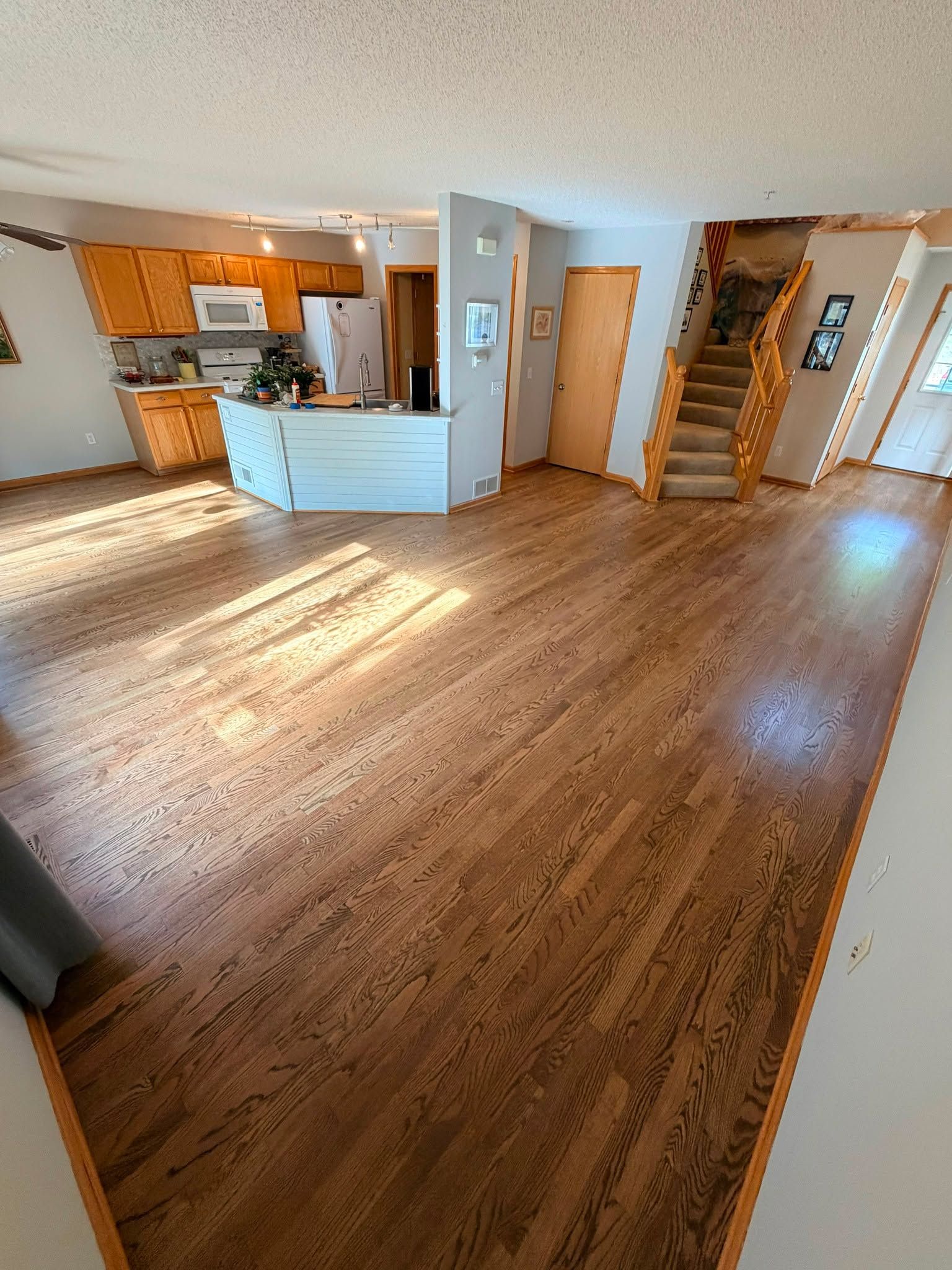 Hardwood plank flooring refinished in medium brown tone throughout open kitchen and living area with wooden cabinetry and stairs visible.
