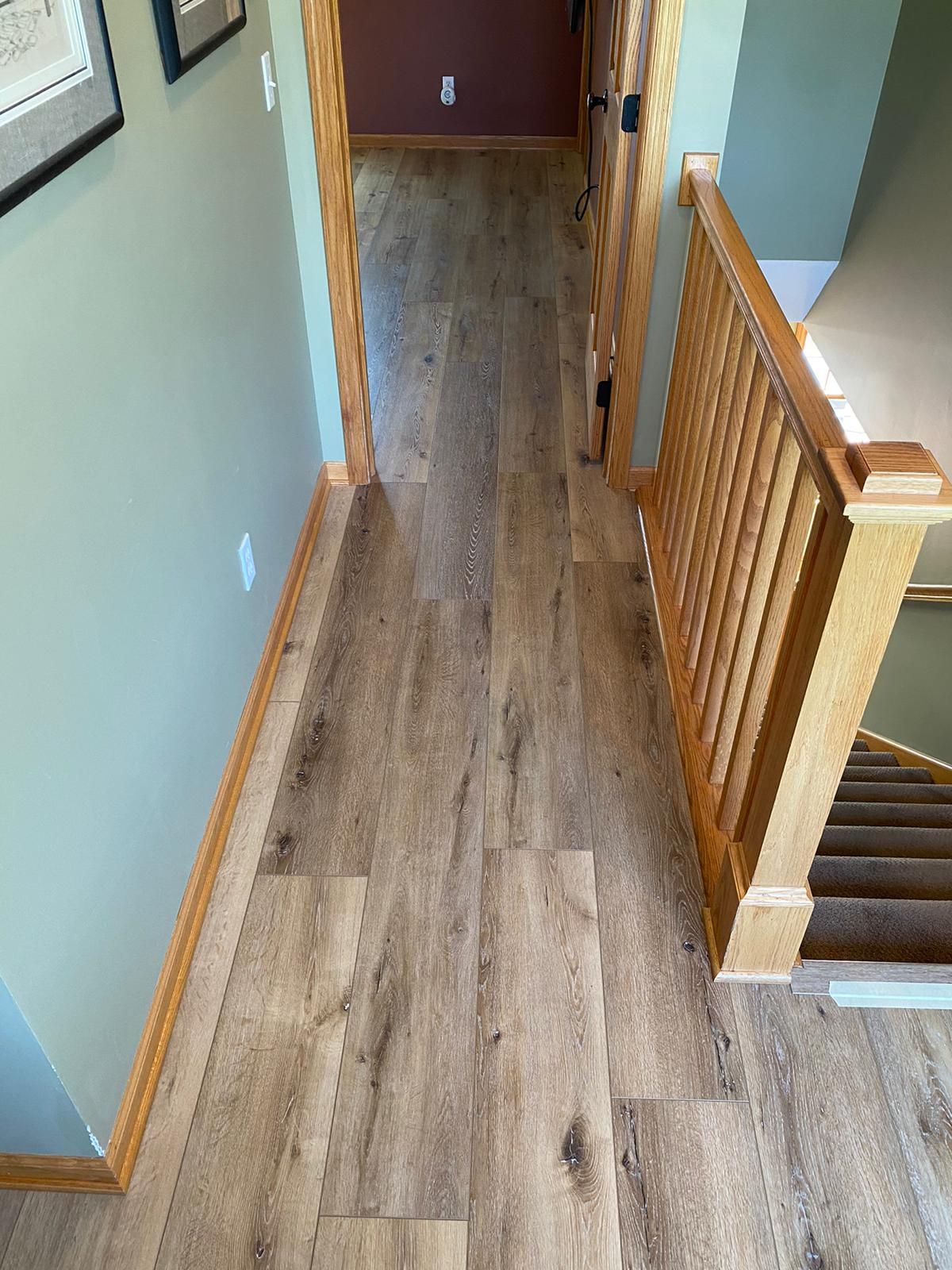 Hallway with medium-toned wood-look LVP planks running lengthwise toward stairs and doorway.