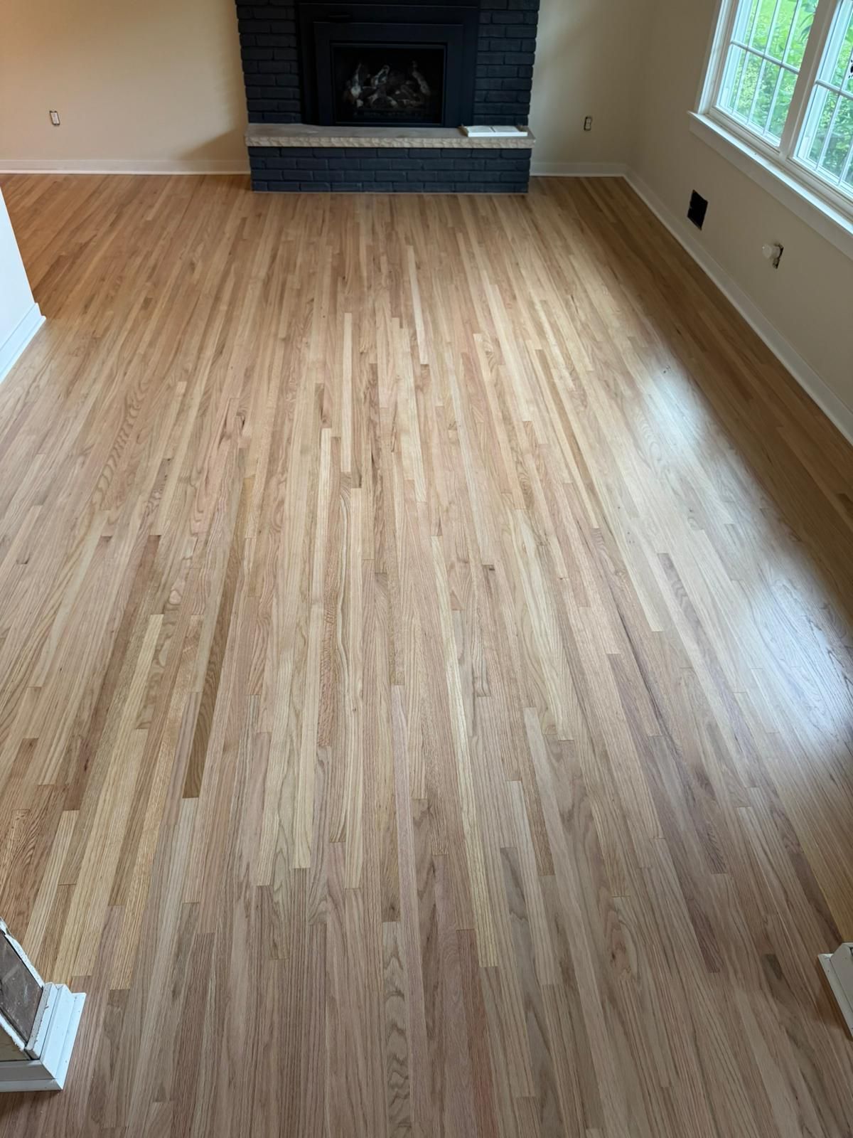 Medium-toned hardwood plank flooring in a living room with a fireplace and windows.