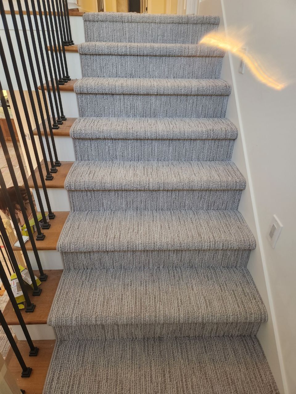 Gray textured carpet runner installed on stairs with wooden treads and black metal railing.