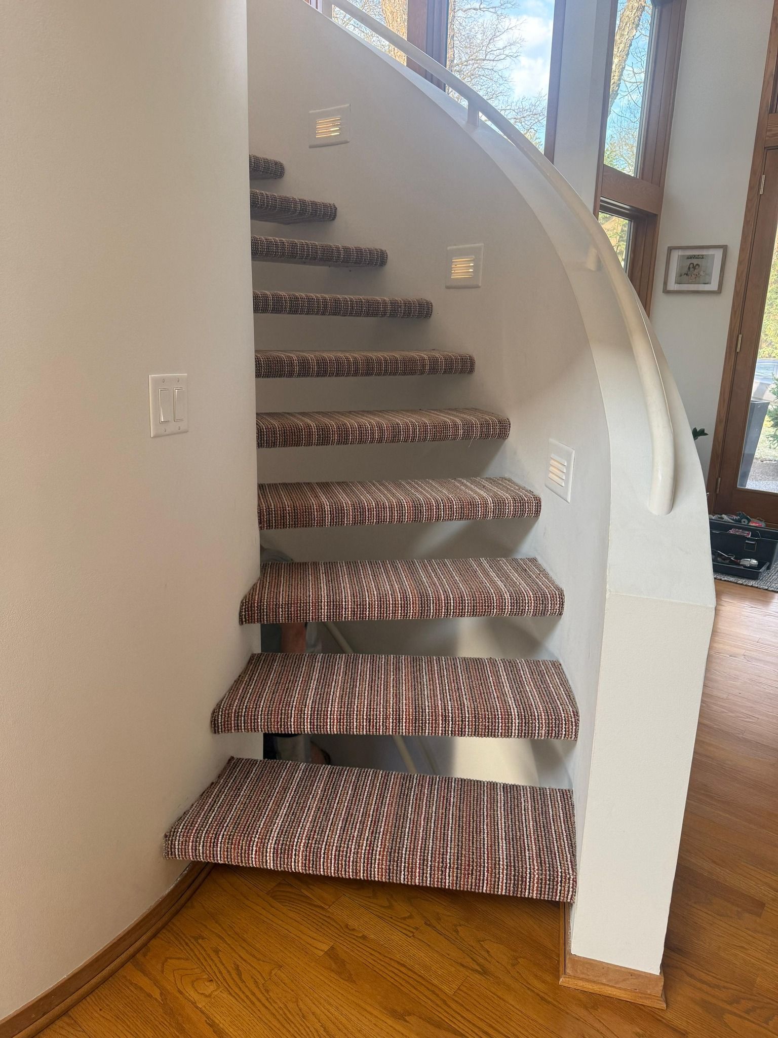 Staircase with brown and red striped carpet treads and metal nosing installed in a curved stairwell with white walls.