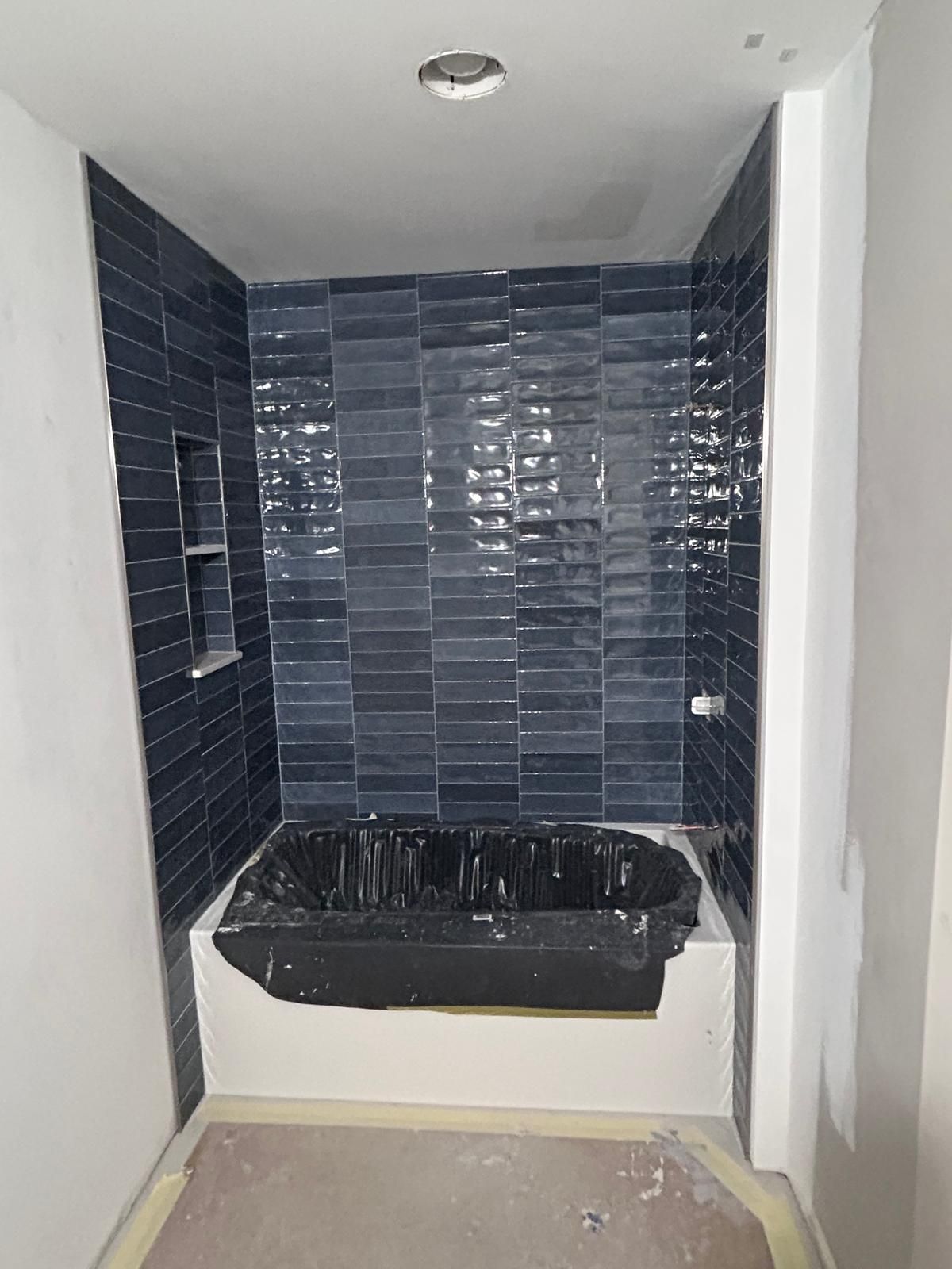 Black subway tile shower surround with white bathtub and gray ceiling in bathroom under construction.