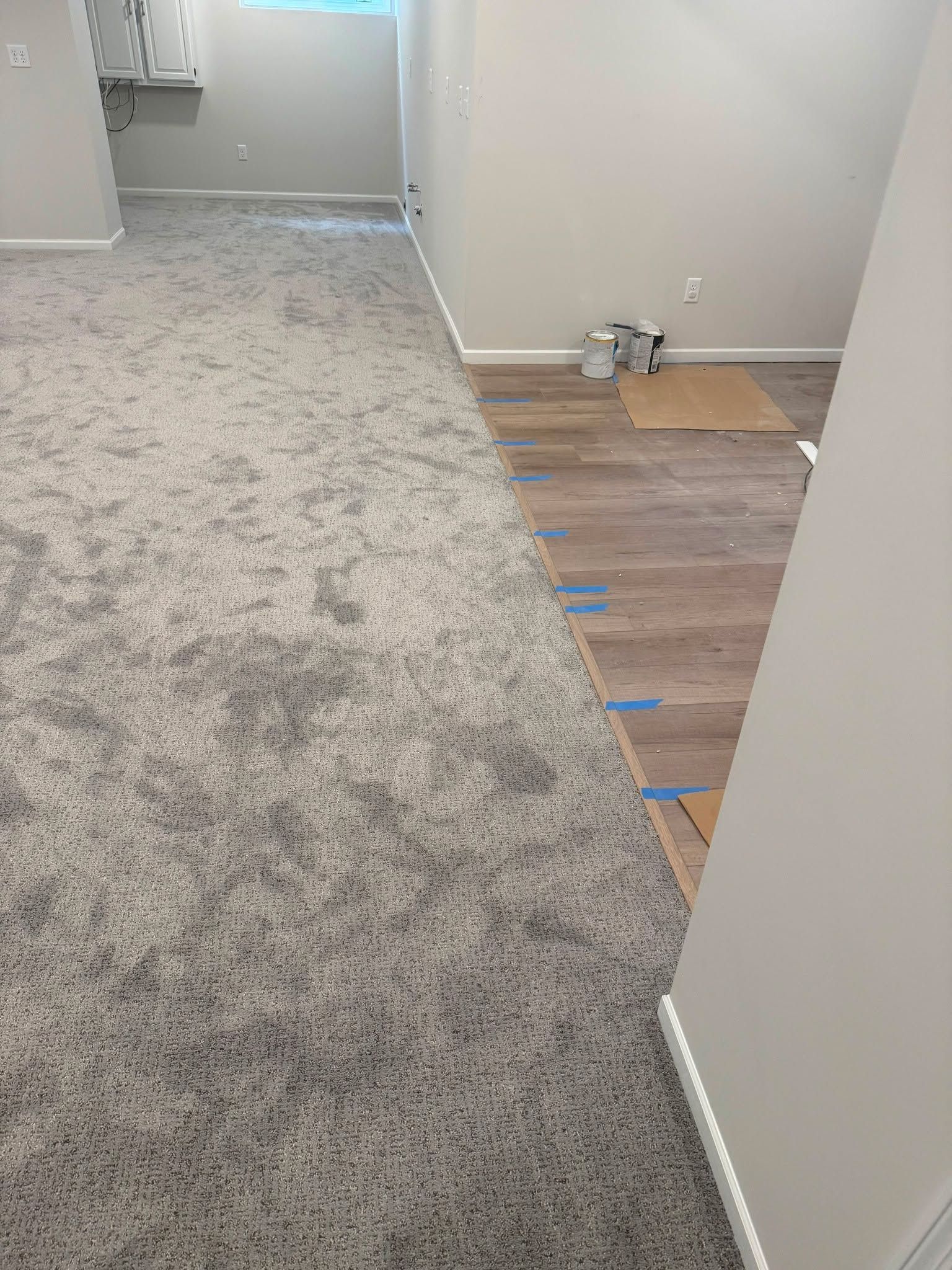Basement room with gray carpet installation in progress next to wood flooring with blue tape marking the transition line.