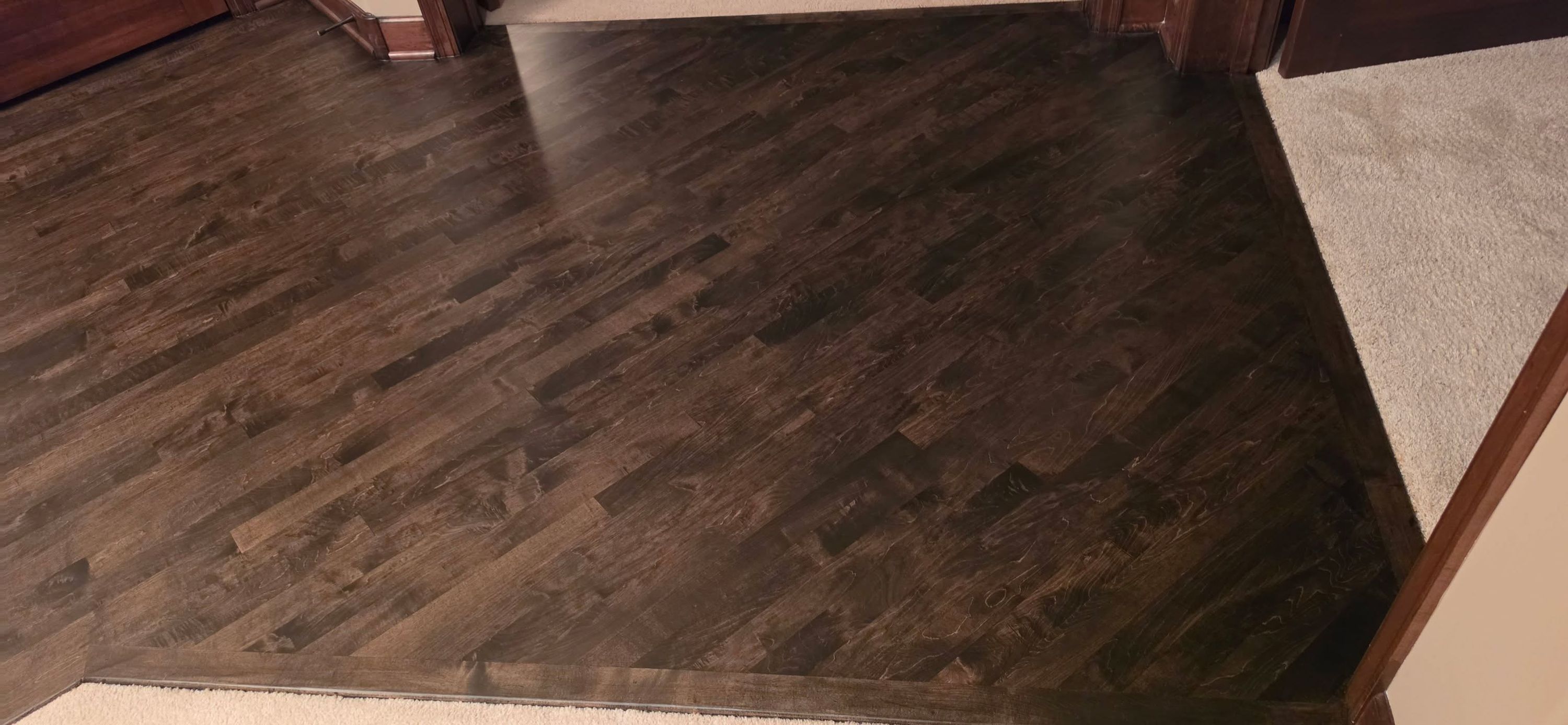 Dark brown maple hardwood planks in living area with carpet transition visible.