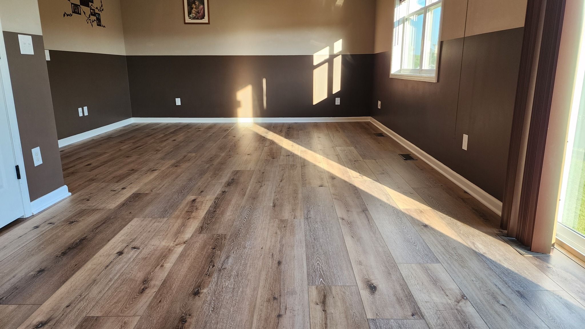 Large living room with medium-toned wood-look LVP planks laid in a staggered pattern, with dark brown upper walls and light upper walls.