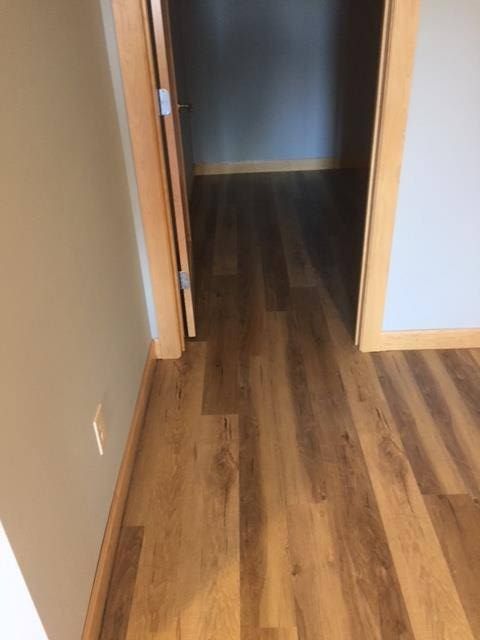 Hallway with medium brown plank-style LVP flooring leading to a dark room with light-colored walls and wooden door frames.