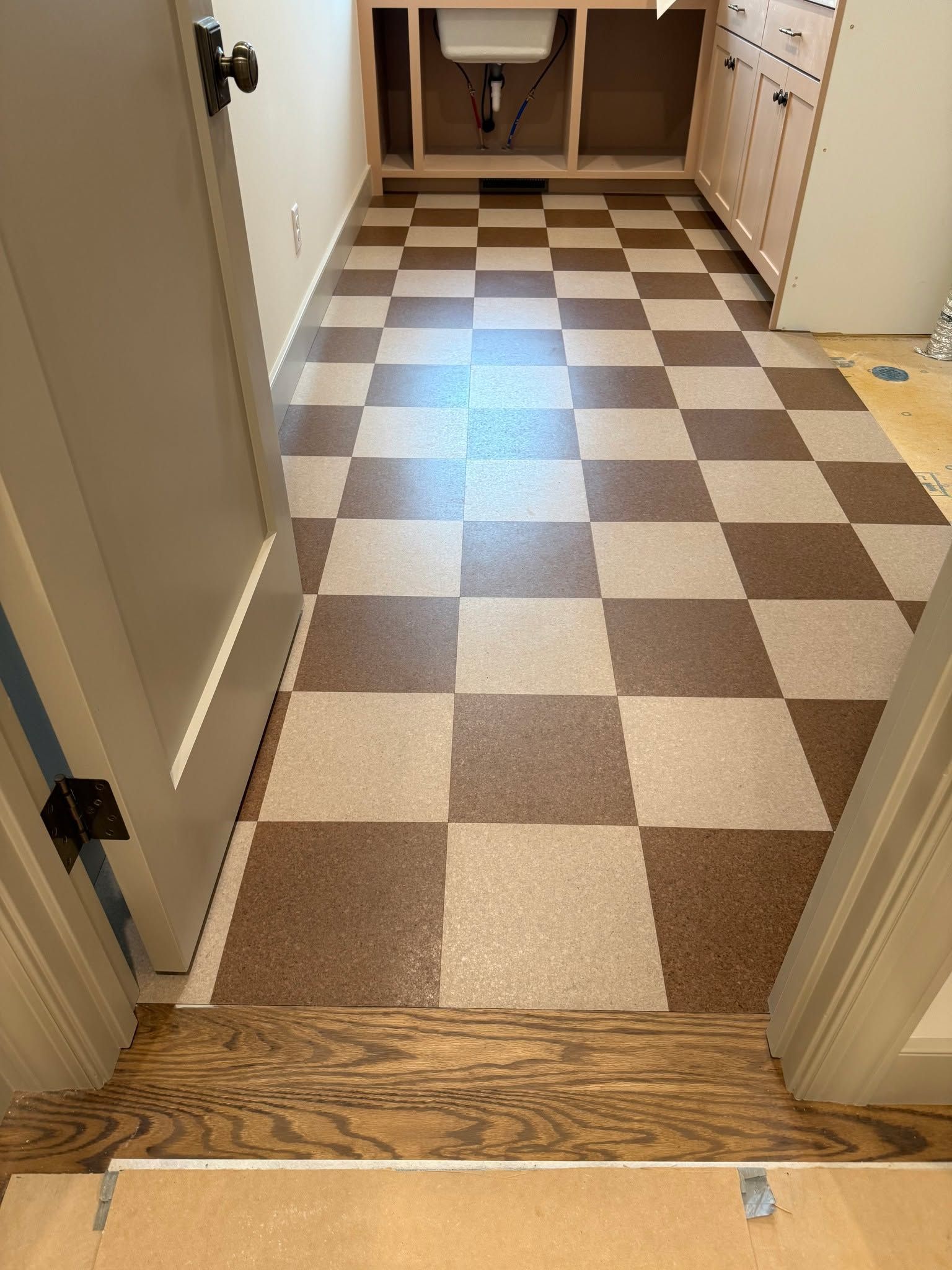 Cork tile floor in checkerboard pattern with tan and brown squares in a laundry room with white cabinetry and hardwood flooring visible in doorway.