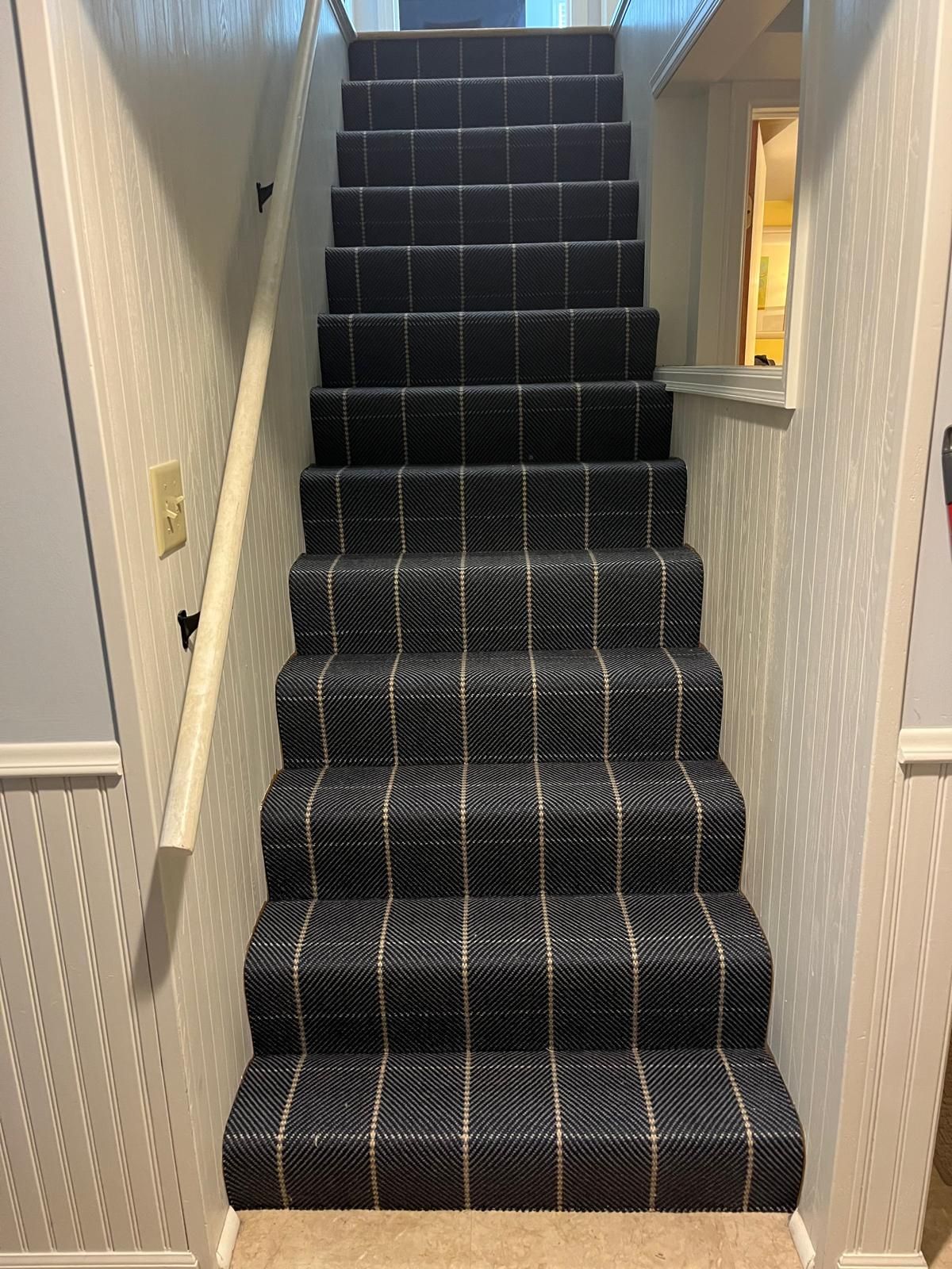 Black carpet with white plaid pattern installed on basement stairs with white painted walls.