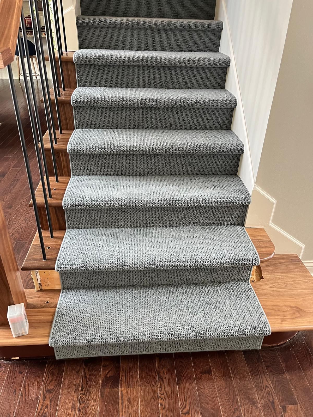 Gray carpet runner installed on wooden stairs with metal railing and white walls.