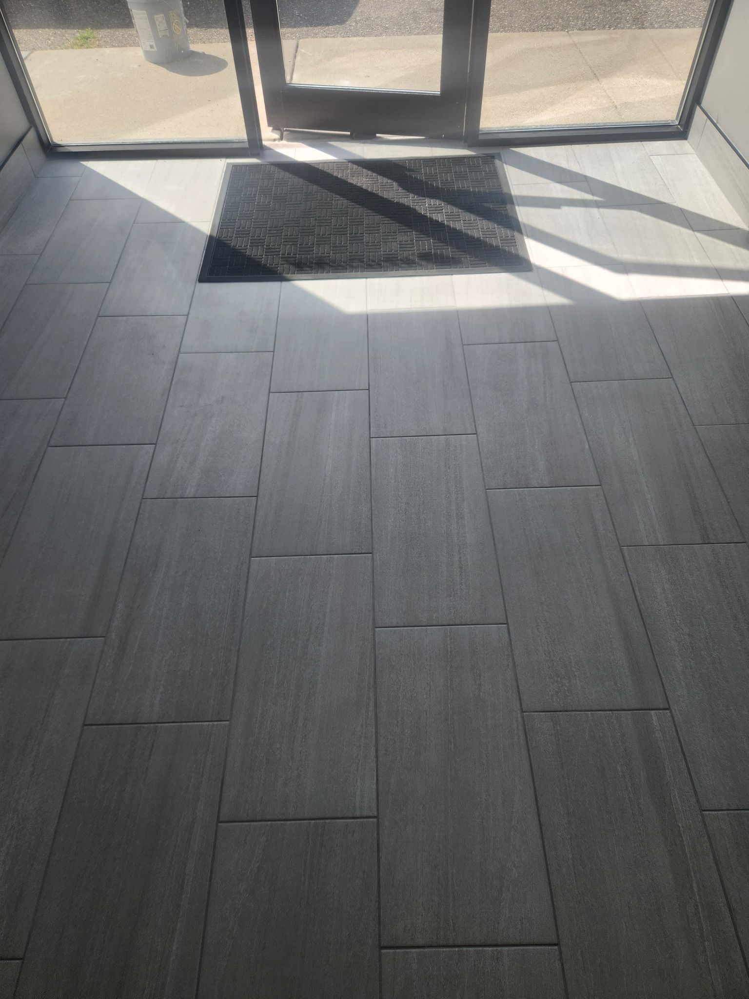 Gray plank-pattern tile flooring in an entry area with a dark mat and glass doors leading outside.