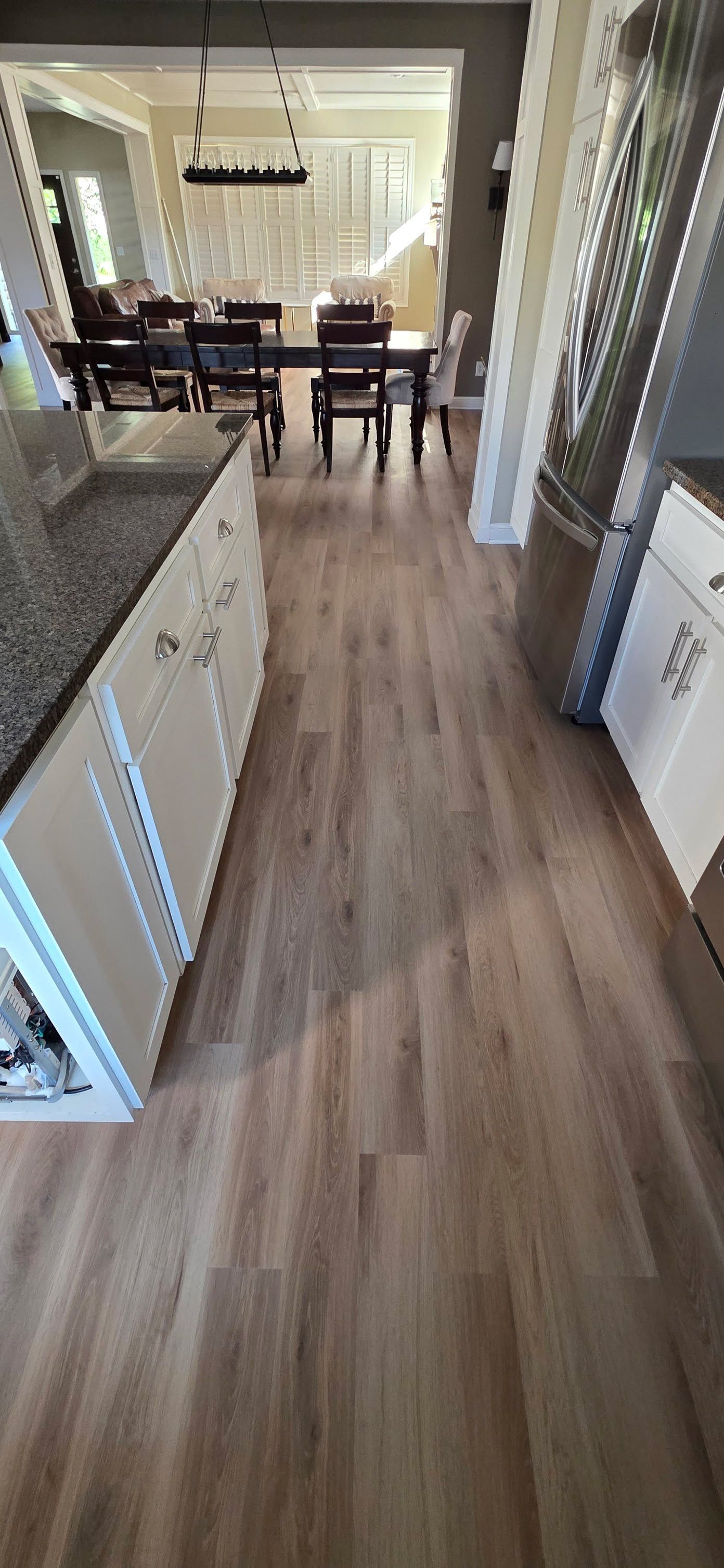 Kitchen with brown plank LVP flooring extending into dining area with white cabinetry and dark dining chairs visible.