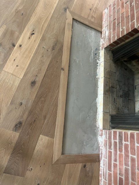 Light tan and brown hardwood plank flooring installed around a fireplace opening with brick surround.